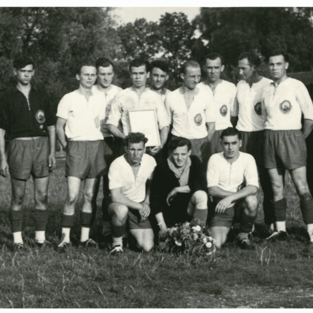 Schwarz-weißes Gruppenbild von Fußballspielern mit Pokal, umgeben von mehreren Männern auf einem Sportplatz.
