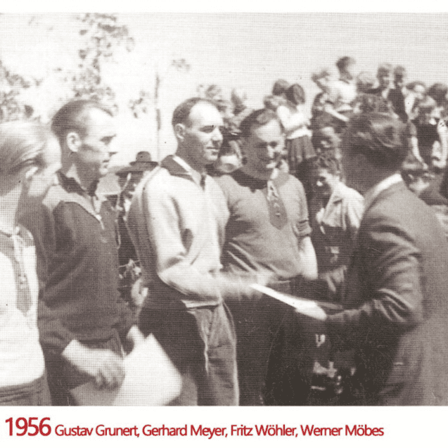 Gruppe von Männern erhält 1956 Urkunden bei einer Ehrung im Freien, Zuschauer im Hintergrund.