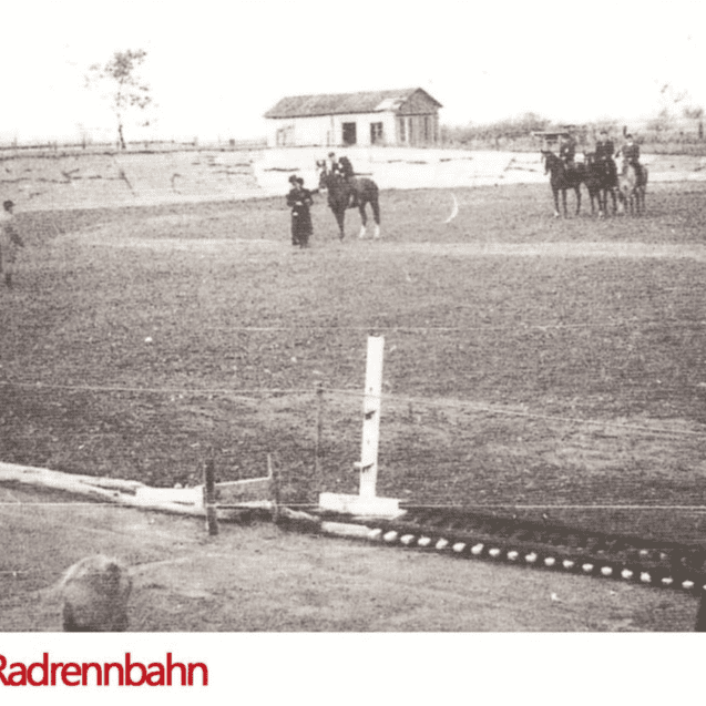 Historisches Foto eines Reitplatzes mit Pferden, Reitern und Zuschauern, im Hintergrund eine kleine Holzscheune.