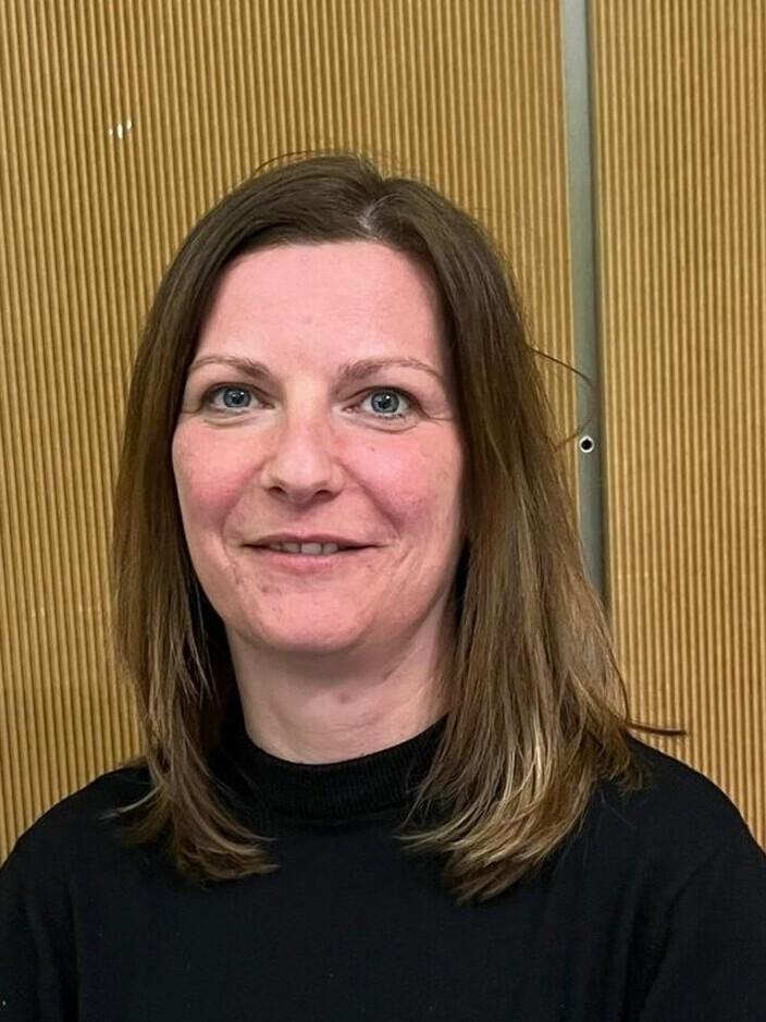 Porträt einer Frau mit langen, braunen Haaren, die vor einer strukturierten Holzvertäfelung steht.