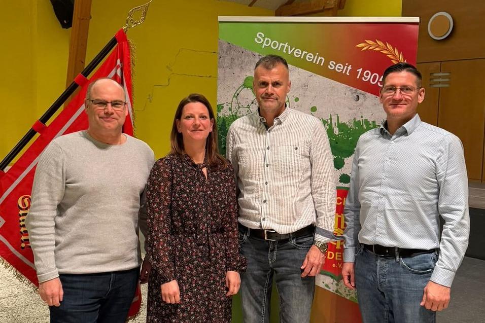 Vier Personen stehen lächelnd vor einem Banner des Sportvereins mit einer roten Fahne im Hintergrund, in einem hellen Raum.