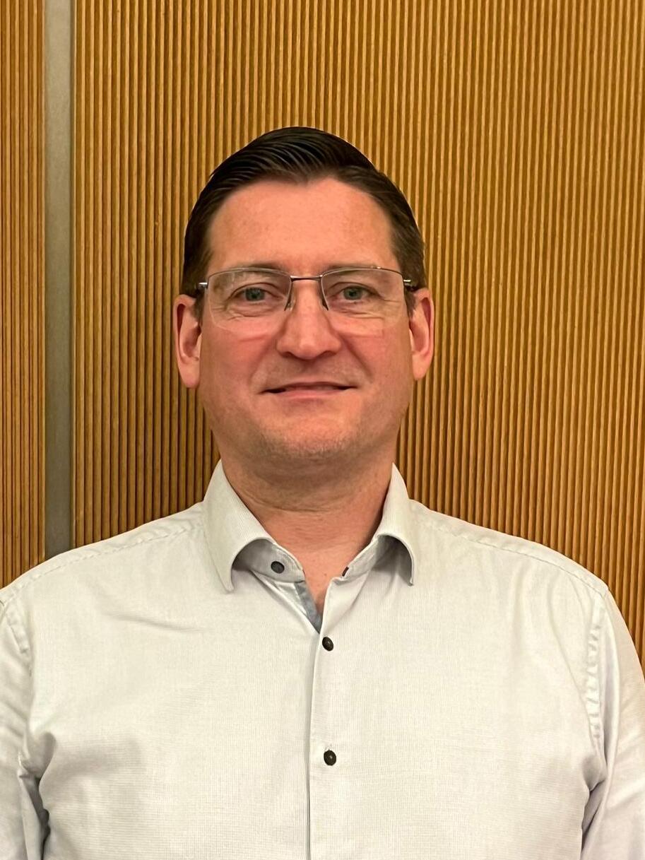 Mann mit Brille in einem hellen Hemd vor einer strukturierten Holzpaneel-Wand.