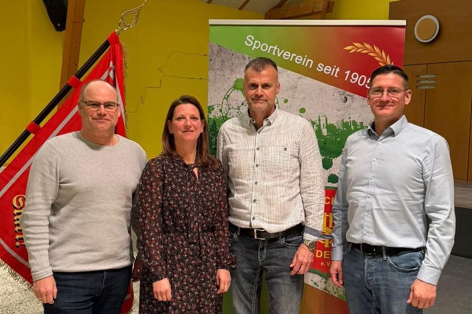 Vier Personen stehen l&auml;chelnd vor einem Banner des Sportvereins mit einer roten Fahne im Hintergrund, in einem hellen Raum.