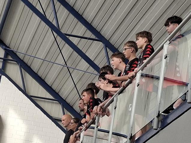 Gruppe junger Menschen in Sportbekleidung steht an einem Balkon und schaut nach unten zu einem Spiel.