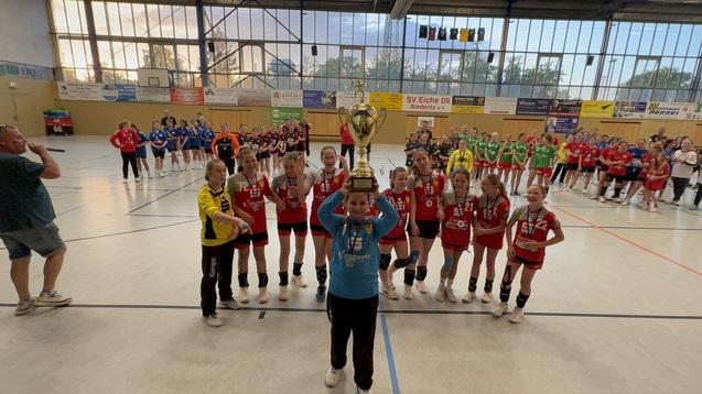Mädchenmannschaft hält einen Pokal in einer Sporthalle, während Zuschauer in verschiedenen Teamtrikots applaudieren.