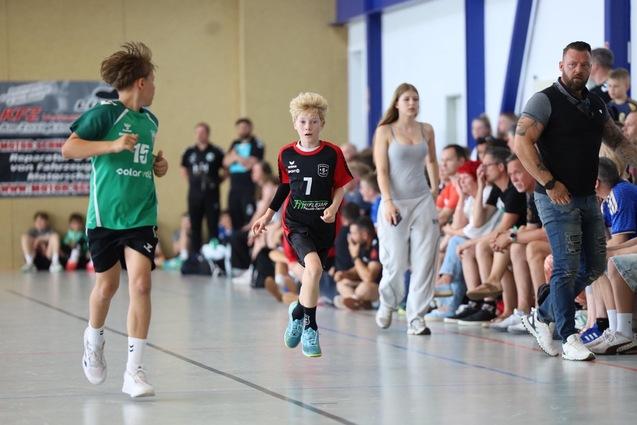 Zwei Kinder im Wettkampf auf einem Handballfeld, umgeben von Zuschauern. Ein Junge in rot-schwarz sprintet, ein anderer trägt grün.