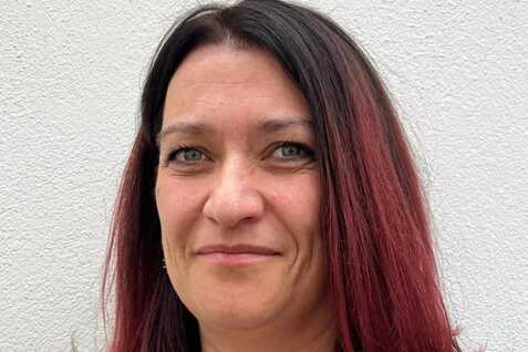 Frau mit schulterlangen, rot-schwarzen Haaren und blauen Augen, tr&auml;gt ein blaues Shirt vor einer wei&szlig;en Wand.
