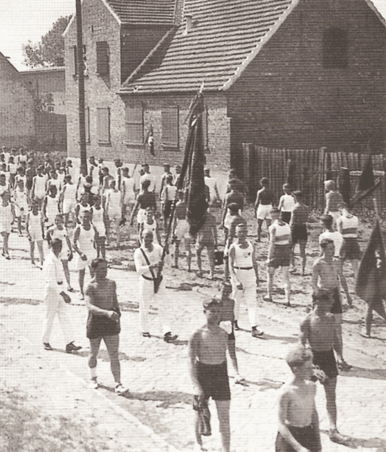 Gruppierung von Männern in Sportbekleidung, die eine Straße entlang marschieren, mit Gebäuden und Fahnen im Hintergrund.