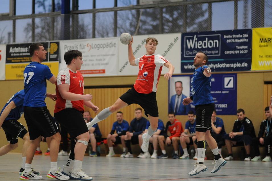 Ein Spieler in rotem Trikot springt zum Wurf, um den Ball w&auml;hrend eines Handballspiels zu werfen, umgeben von Gegnern.