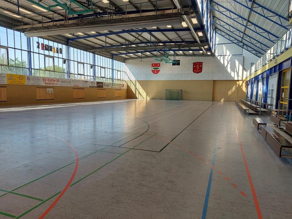 Leere Sporthalle mit Holzfußboden, großen Fenstern und zwei kleinen Toren an der Wand.