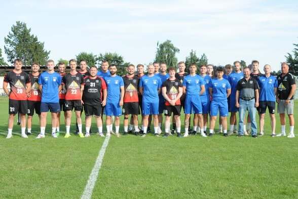 Gruppenfoto einer Fu&szlig;ballmannschaft mit 24 Spielern und Trainern auf einem Rasenplatz unter blauen Himmel.