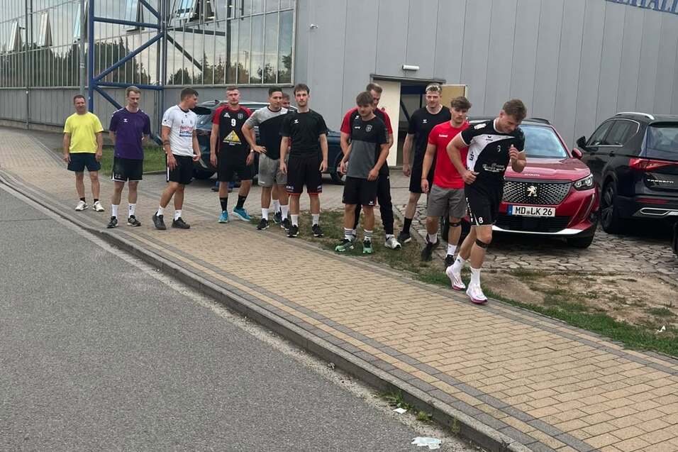 Gruppe von M&auml;nnern in Sportkleidung vor dem Biederitzer Ehlert-Haus, mit Autos und einem Gehweg im Hintergrund.