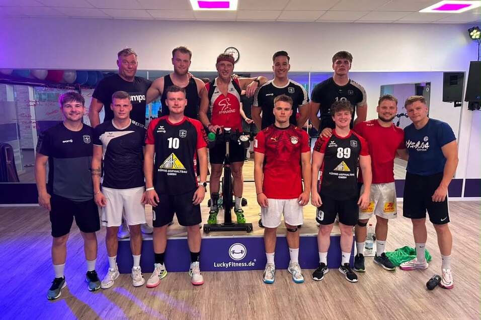 Gruppe von zw&ouml;lf M&auml;nnern in Sportkleidung, die l&auml;cheln und auf einer B&uuml;hne in einem Fitnessstudio posieren.