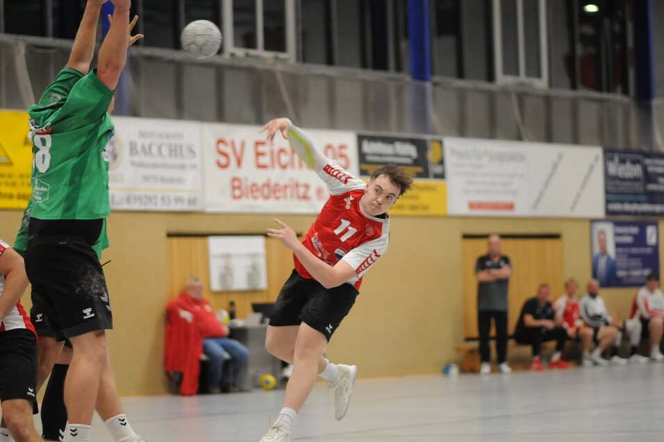 Handballspieler in roter Uniform wirft den Ball w&auml;hrend eines Spiels, der Torwart in gr&uuml;ner Uniform reagiert.