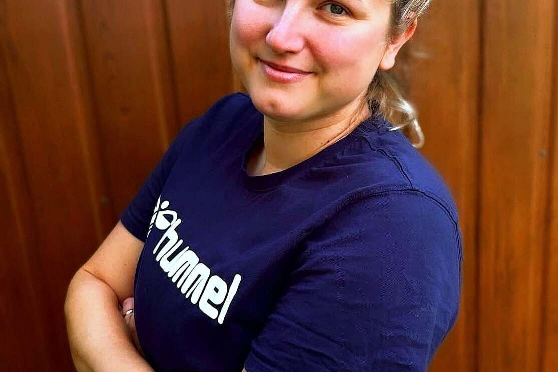 Porträt einer lächelnden Frau mit zusammengesteckten Haaren, die vor einer Holzvertäfelung steht. Sie trägt ein dunkelblaues T-Shirt.