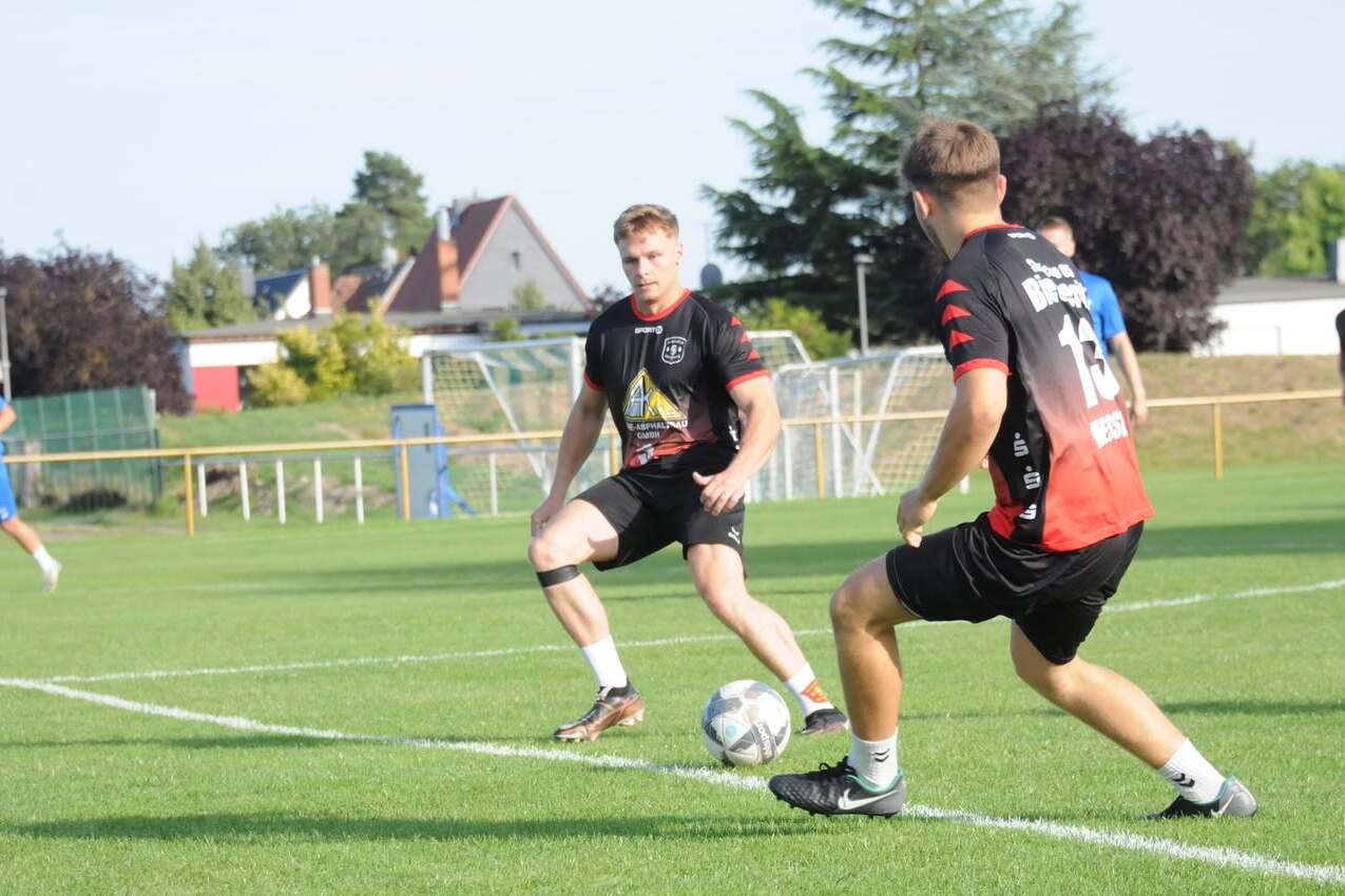 Zwei Fußballspieler im Training passen einen Ball auf einem grünen Rasenplatz unter blauem Himmel.