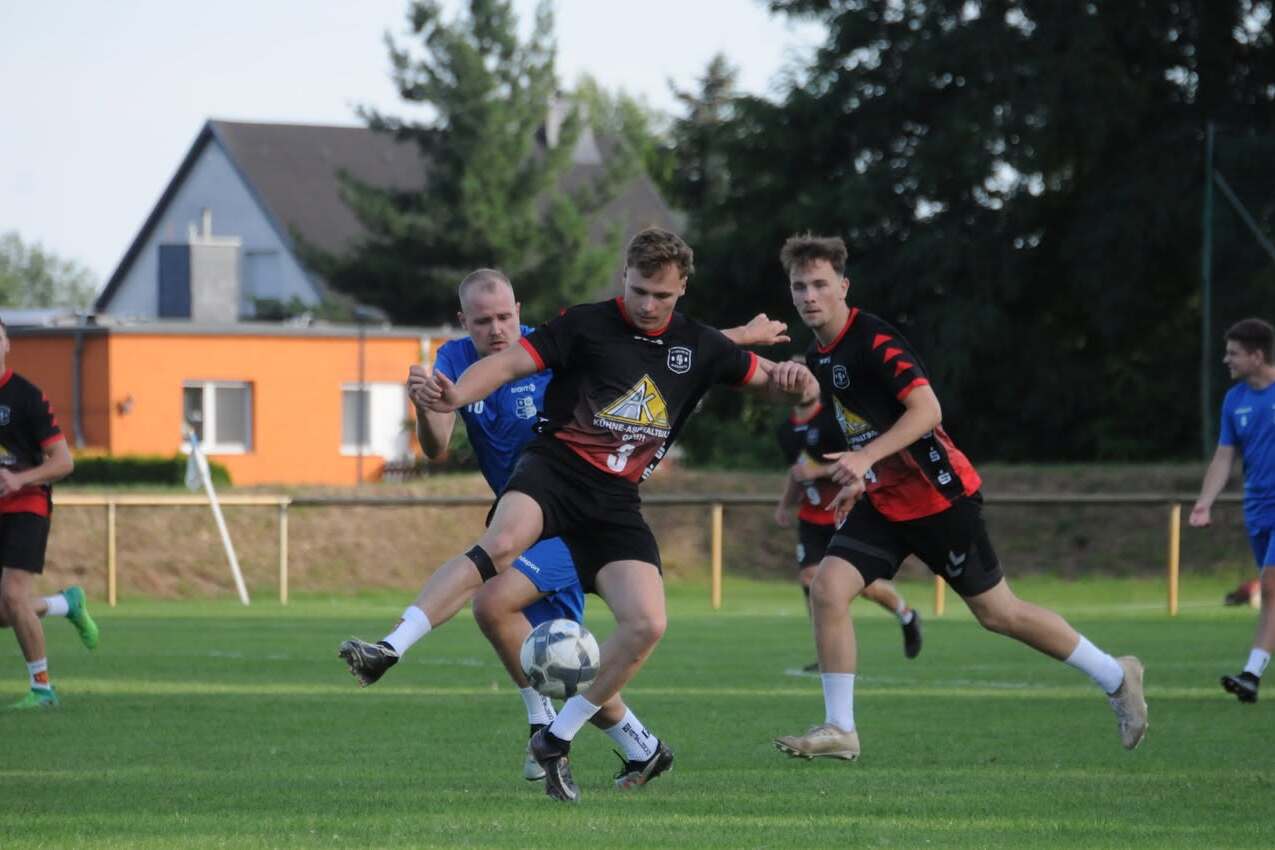 Zwei Fußballspieler kämpfen um den Ball, während ein dritter Spieler im Hintergrund zuschaut, auf einem grünen Spielfeld.
