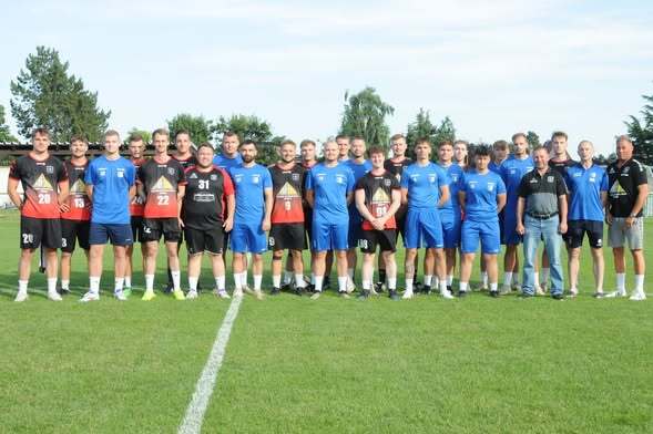 Gruppenfoto einer Fu&szlig;ballmannschaft mit 24 Spielern und Trainern auf einem Rasenplatz unter blauen Himmel.