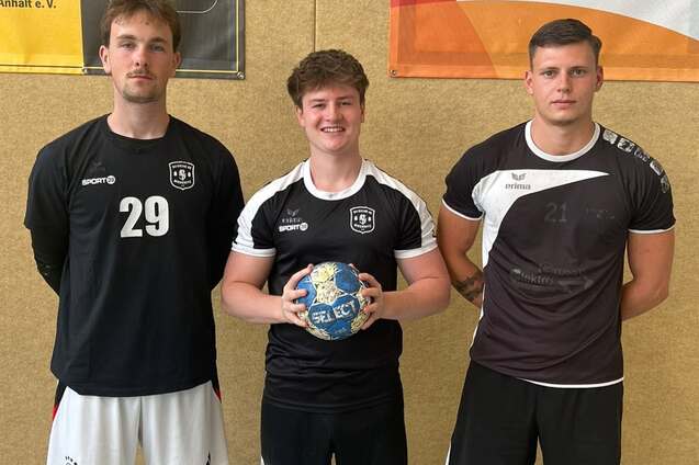 Drei Handballspieler stehen vor einer Wand mit Sportplakaten und halten einen Handball in der Mitte.