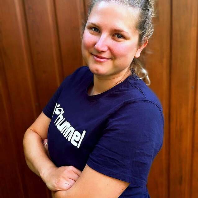 Porträt einer lächelnden Frau mit zusammengesteckten Haaren, die vor einer Holzvertäfelung steht. Sie trägt ein dunkelblaues T-Shirt.