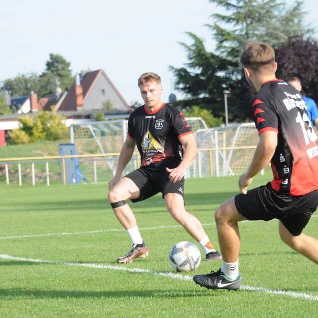 Zwei Fußballspieler im Training passen einen Ball auf einem grünen Rasenplatz unter blauem Himmel.
