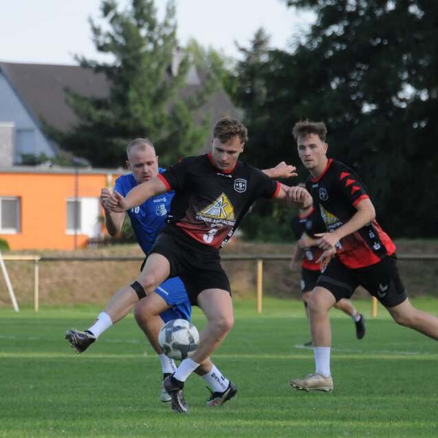 Zwei Fußballspieler kämpfen um den Ball, während ein dritter Spieler im Hintergrund zuschaut, auf einem grünen Spielfeld.