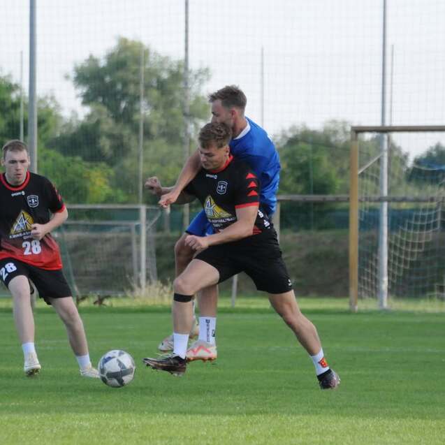 Gruppen von Fußballspielern auf einem Rasenfeld, ein Spieler wird von hinten am Ball gehindert.