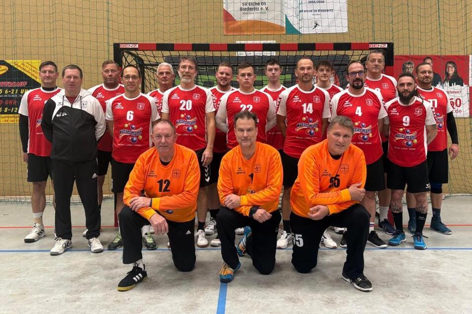Mannschaftsaufstellung von 14 Spielern in roten Trikots mit zwei Torh&uuml;tern in orange vor einer Sporthalle.