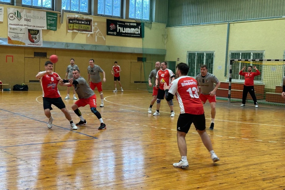 Handballspiel in einer Sporthalle mit Spielern in roten und grauen Trikots, die sich um den Ball bewegen.