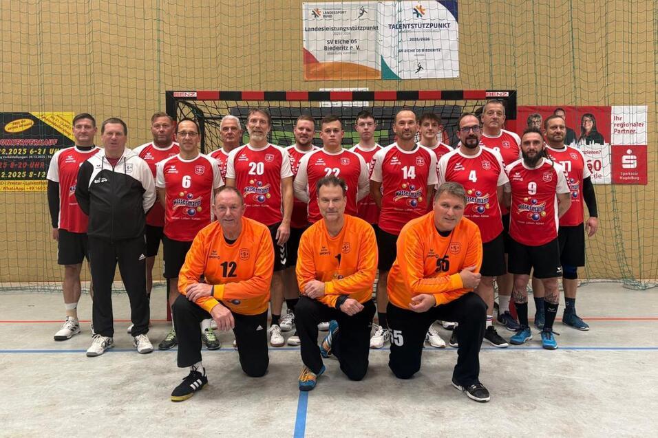 Mannschaftsaufstellung von 14 Spielern in roten Trikots mit zwei Torh&uuml;tern in orange vor einer Sporthalle.