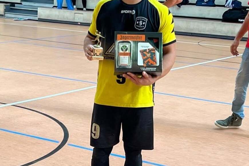 Ein junger Mann in Sportkleidung h&auml;lt einen Pokal und eine Auszeichnung in einer Sporthalle.