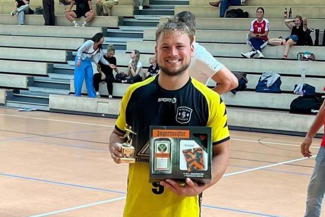 Ein junger Mann in Sportkleidung h&auml;lt einen Pokal und eine Auszeichnung in einer Sporthalle.