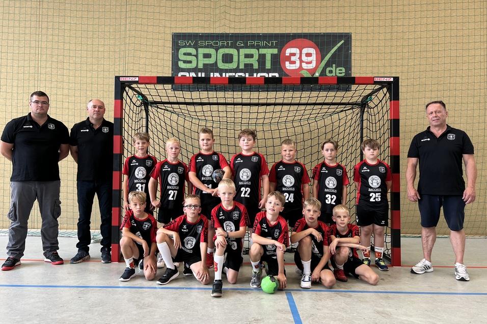 Gruppe junger Handballspieler in Sportkleidung steht vor einem Tor auf einem Hallenboden, begleitet von zwei Trainern.