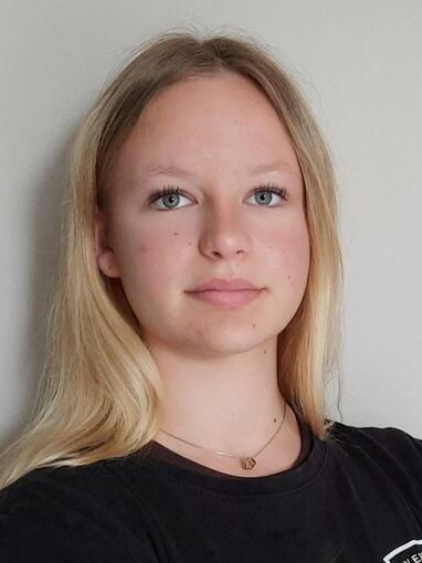 Portr&auml;t einer jungen Frau mit langen, blonden Haaren, die vor einer hellen Wand steht und direkt in die Kamera schaut.