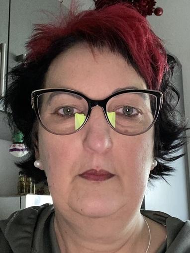 Portr&auml;t einer Frau mit rot-schwarzen Haaren, Brille und gr&uuml;nen Akzenten am Brillenrahmen, vor klarer Hintergrund.