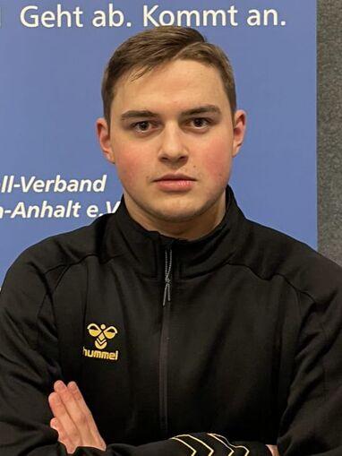 Junger Mann in schwarzer Sportjacke mit crossed arms, vor einer blauen Hintergrundwand mit Text.