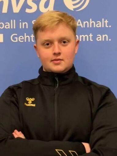 Portr&auml;t eines jungen Mannes in schwarzer Jacke mit verschr&auml;nkten Armen vor einem blauen Hintergrund mit Handball-Logo.