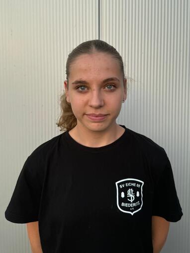 Portr&auml;t einer jungen Frau mit Pferdeschwanz, getragenem schwarzem T-Shirt mit Logo, vor einer strukturierten, hellen Wand.