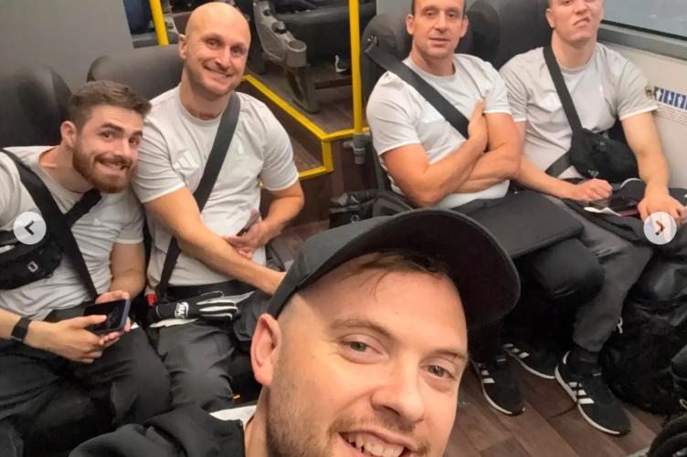 Fünf Männer in Sportkleidung sitzen im Bus und schauen in die Kamera. Der vordere Mann lächelt.