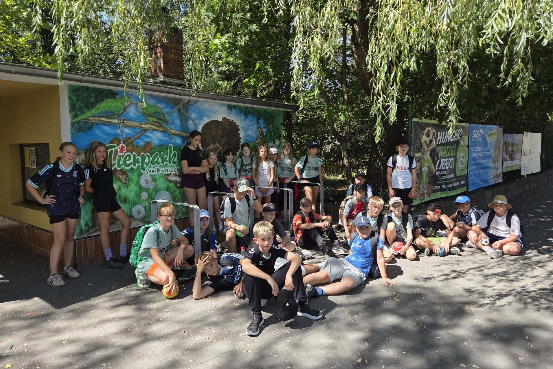 Gruppierung von Kindern und Jugendlichen, die vor einem bemalten Gebäude mit Naturmotiven und Werbebannern posieren.
