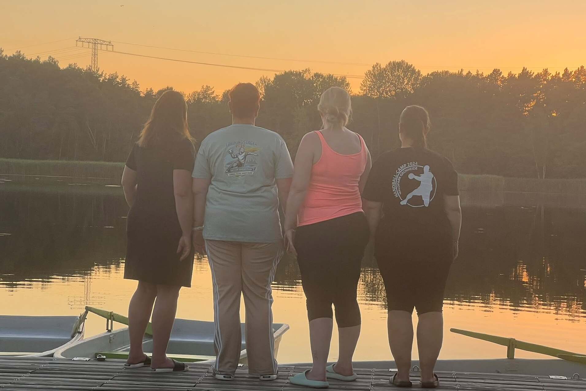 Vier Frauen stehen mit dem Rücken zu einem ruhigen See bei Sonnenuntergang auf einer Holzplattform.