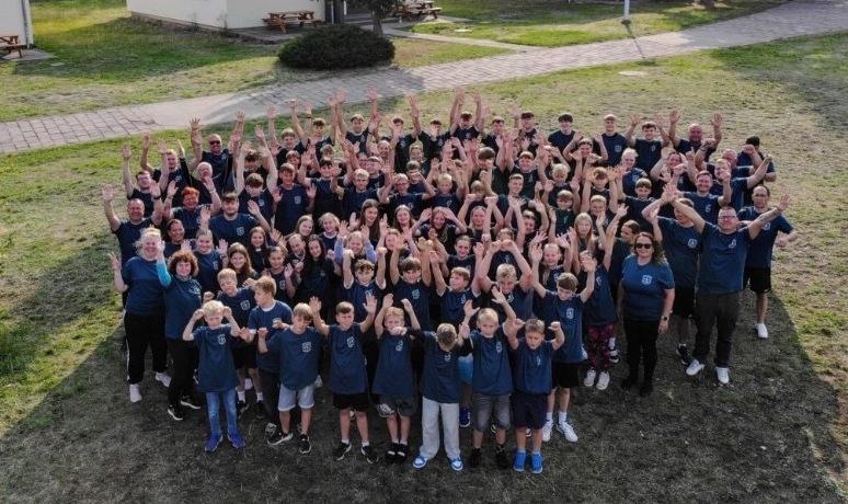 Gruppenfoto von &uuml;ber 150 Personen in blauen T-Shirts, die in einem Freiluftbereich jubeln. Im Hintergrund sind H&uuml;tten sichtbar.