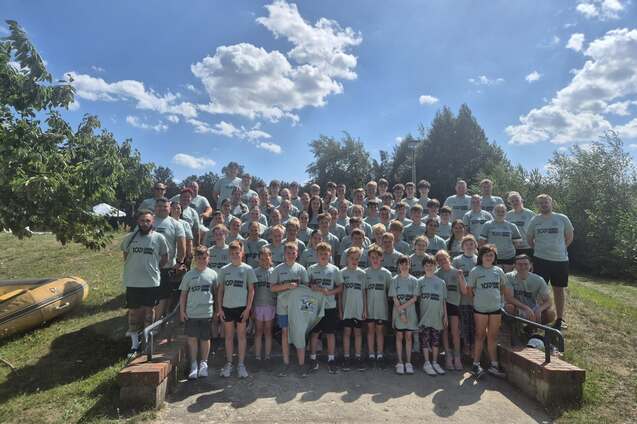 Gruppenfoto von rund 70 Personen in grauen T-Shirts, umgeben von B&auml;umen und blauem Himmel mit Wolken.