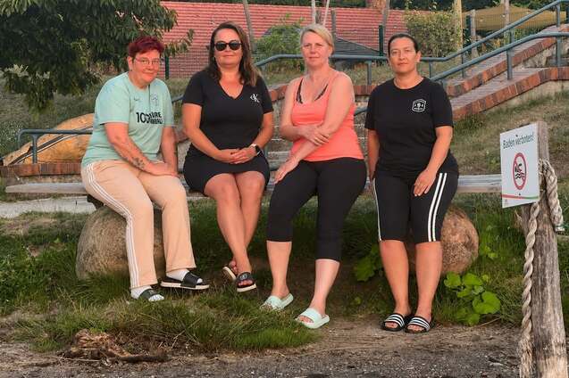 Vier Frauen sitzen auf Baumst&auml;mmen am Ufer eines Gew&auml;ssers, umgeben von Pflanzen und einer Holztreppe im Hintergrund.