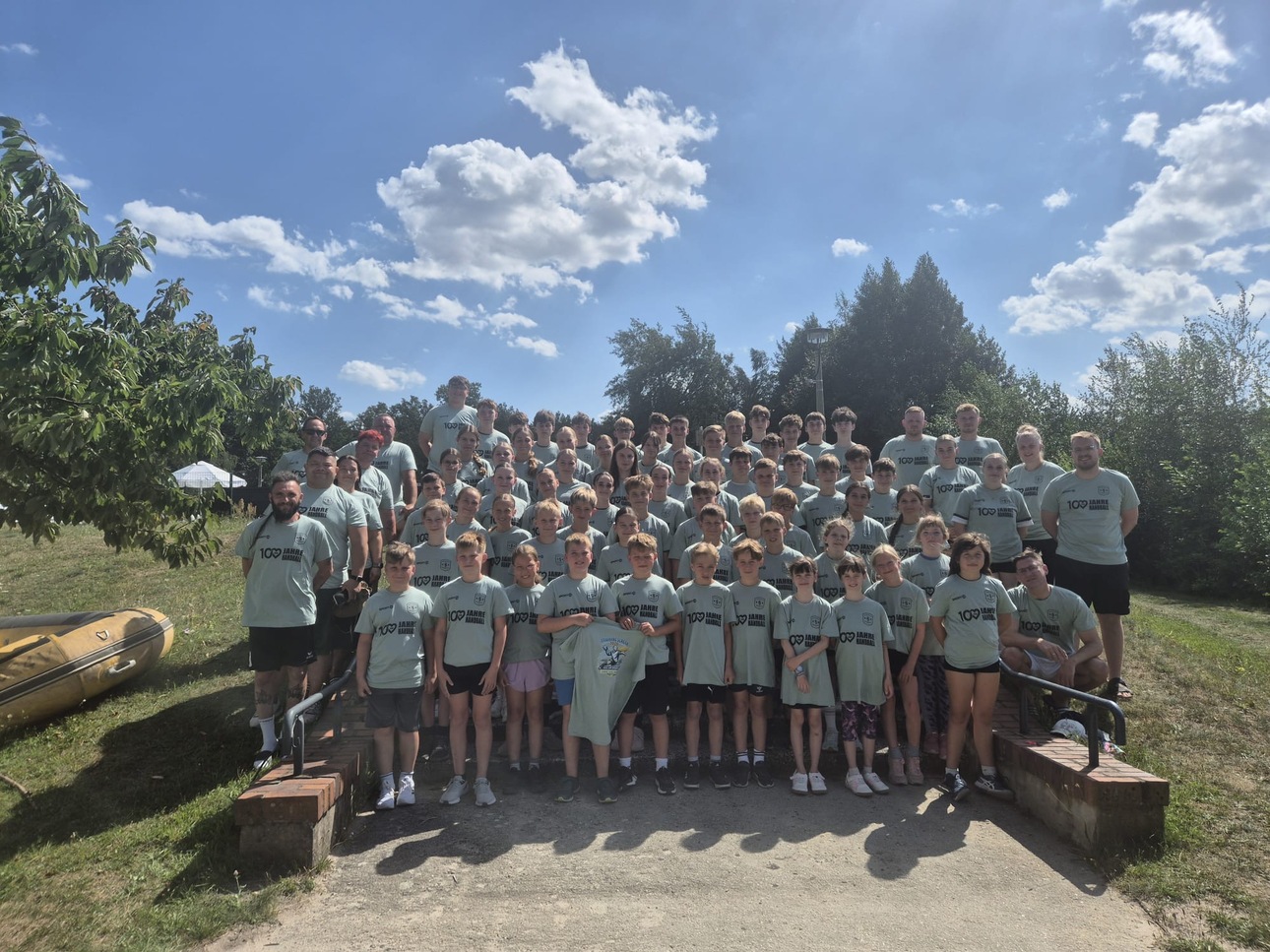 Gruppenbild von &uuml;ber 100 Personen in gr&uuml;nen T-Shirts, stehend auf einer Wiese unter blauem Himmel mit Wolken.