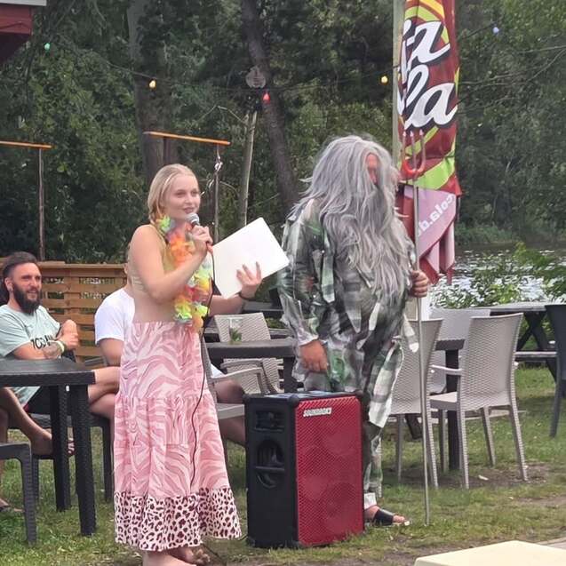 Frau mit Lei-Blumenkranz spricht am Mikrofon, während ein Mann mit grauen Haaren daneben steht, Pool im Hintergrund.