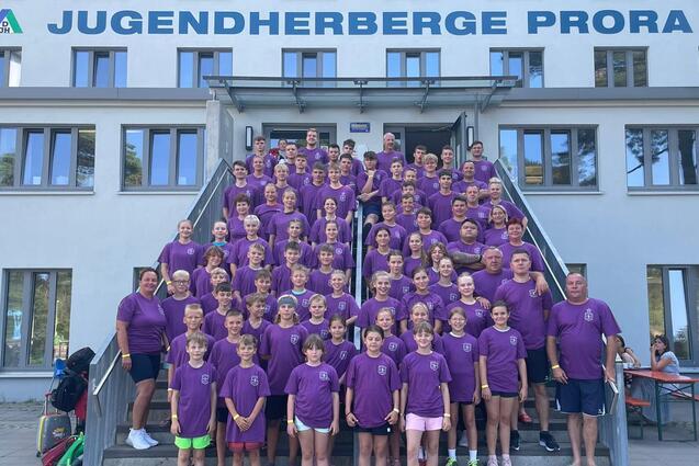 Gruppenfoto von &uuml;ber 100 Personen in lila T-Shirts vor der Jugendherberge Prora, auf Treppe und im Hintergrund.
