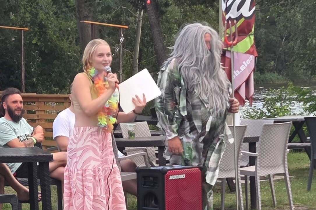 Frau mit Lei-Blumenkranz spricht am Mikrofon, während ein Mann mit grauen Haaren daneben steht, Pool im Hintergrund.