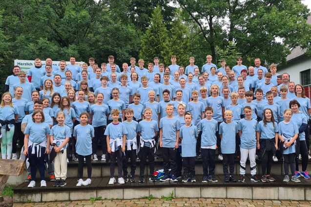 Gruppenfoto von &uuml;ber 100 Personen in blauen T-Shirts, die auf einer Treppe vor einem Geb&auml;ude stehen, umgeben von B&auml;umen.