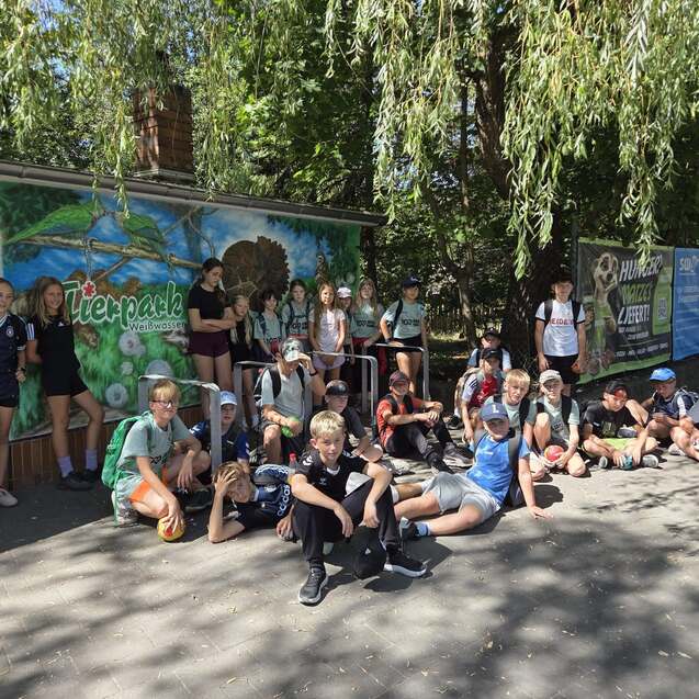 Gruppierung von Kindern und Jugendlichen, die vor einem bemalten Gebäude mit Naturmotiven und Werbebannern posieren.